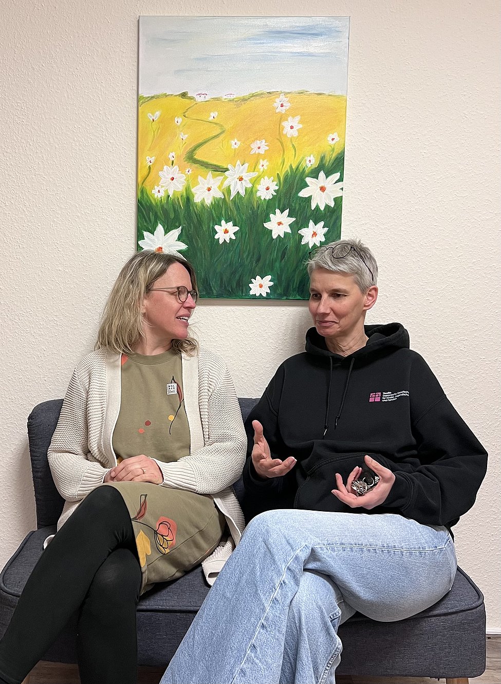 Zwei Frauen sitzen plaudernd auf einem Sofa vor einem bunten Blumenbild und wirken dabei gelöst und freundlich.
