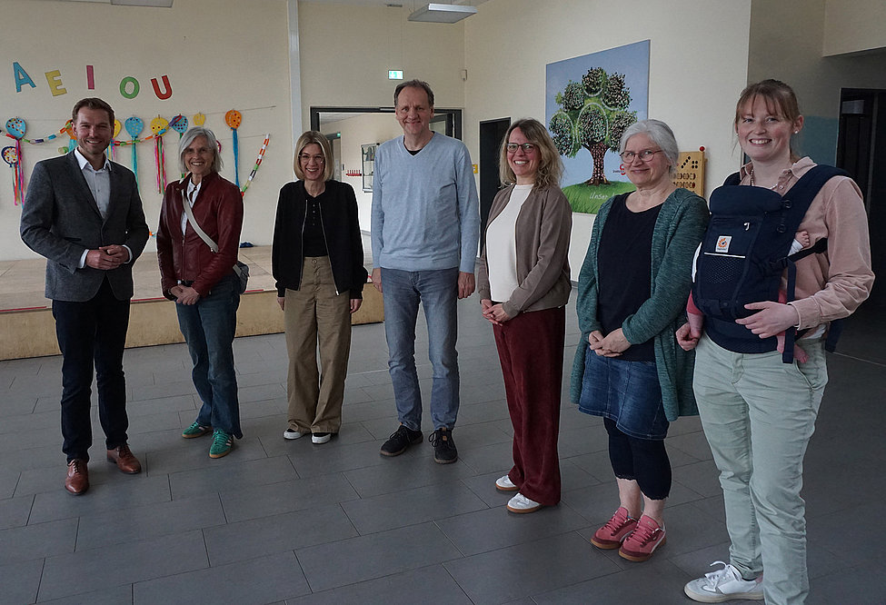Das Foto zeigt sieben Personen, die in einem Halbkreis nebeneinanderstehen. Alle schauen in die Kamera. Im Hintergrund sind Bilder und Gebasteltes zu sehen. 