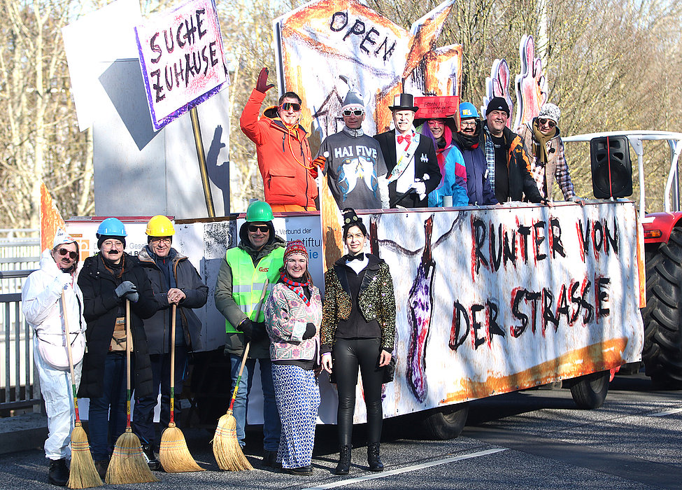 Eine Gruppe verkleideter Menschen steht lächelnd vor einem bunt dekorierten Umzugswagen mit Schildern wie „Suche Zuhause“ und „Runter von der Straße“. 